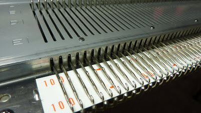 Full-length view of a knitting machine's needlebed showing the fixed number of needles available, illustrating the width limitations machine knitters face when creating larger pieces