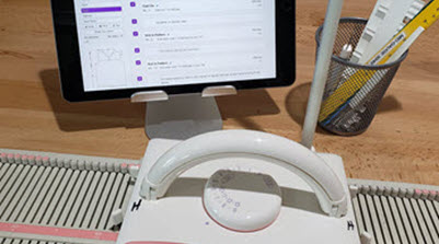 Organized machine knitting workspace showing a knitting machine, tablet with digital pattern, and essential tools ready for project planning