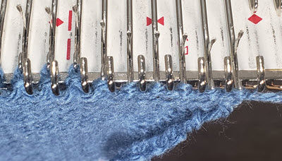 Close-up view of knitting still on the machine bed, showing distorted and overly tight stitches along the edge where the carriage enters, a common issue faced by machine knitters