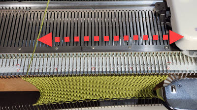 A knitting machine carriage positioned on the opposite side of the working yarn, illustrating the common situation that occurs after ripping out rows or using a garter bar