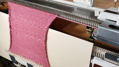 A knitting machine ribber attachment protected by a custom cover, with knitted fabric draped safely over it, demonstrating proper needle bed protection