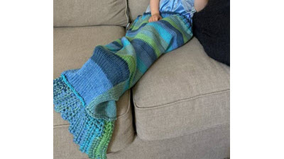 A toddler snuggled inside a knitted mermaid tail blanket with textured stitch patterns, showing the practical use of this cozy children's accessory