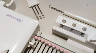 A Silver Reed LK150 knitting machine displayed with essential knitting tools and accessories, including transfer tools and a row counter