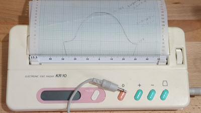 KR10 charting device in use, showing a knitting pattern being traced with a stylus and graph paper for precision machine knitting