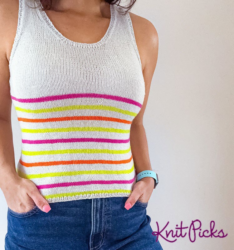 A person wearing a white sleeveless tank with bright pink, lime, and orange horizontal stripes, hands in the pockets of denim shorts.