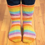A pair of colorful self-striping socks in yellow, orange, pink, purple, and green stripes, worn standing on a wood floor.