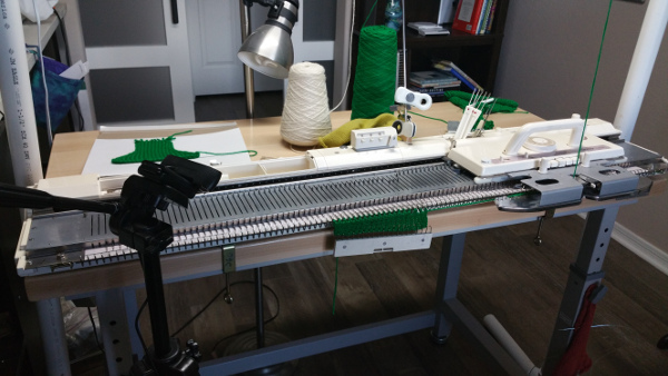 Bulky knitting machine with green knitting on the needles, clamped to a wood-top height-adjustable workbench; yarn cones, a desk lamp, and small tools sit behind the machine.